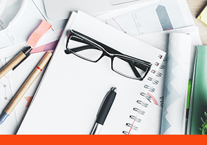 Glasses, pad of paper and pen on desk scattered with office supplies