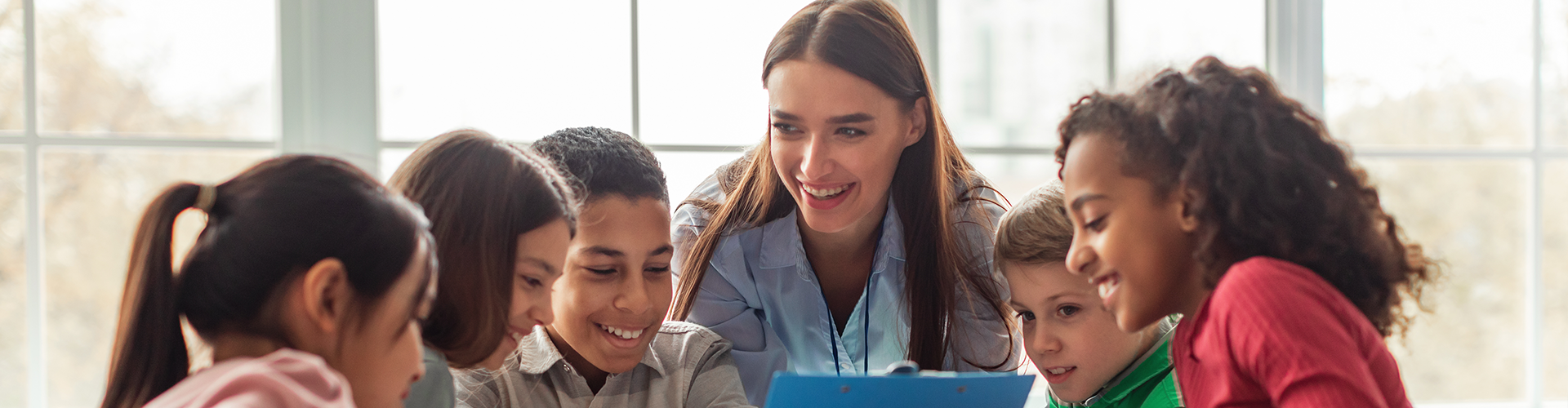 Teacher and five elementary students gathered closely, gazing down with happy expressions at something out of the photo frame