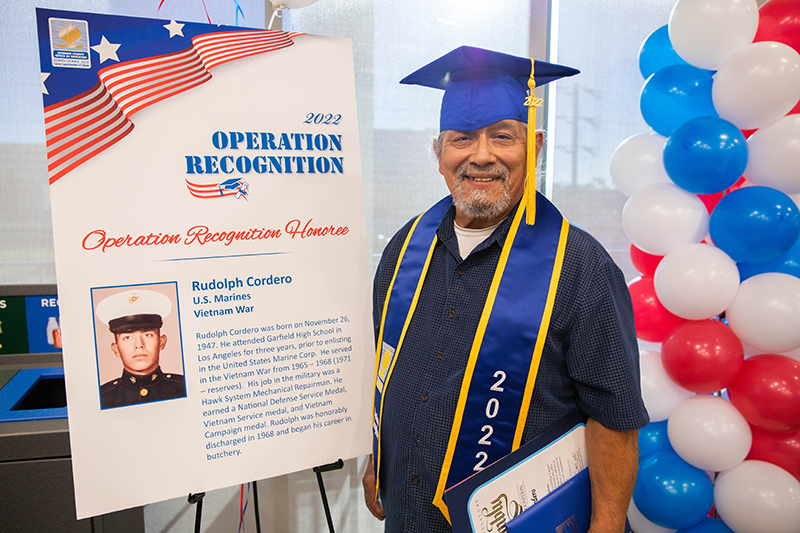 Graduate in cap and gown standing next to an Operation Recognition poster with his military photo and bio