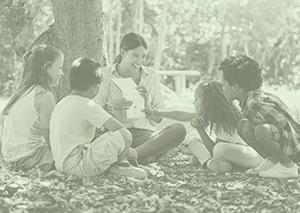 Woman sitting under tree with 4 attentive elementary aged students